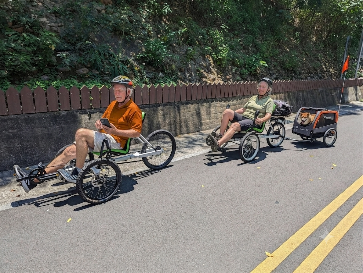 Two senior cyclists ride OVELOX recumbent trikes on a sunny US trail, one towing a pet trailer with a Shiba Inu dog